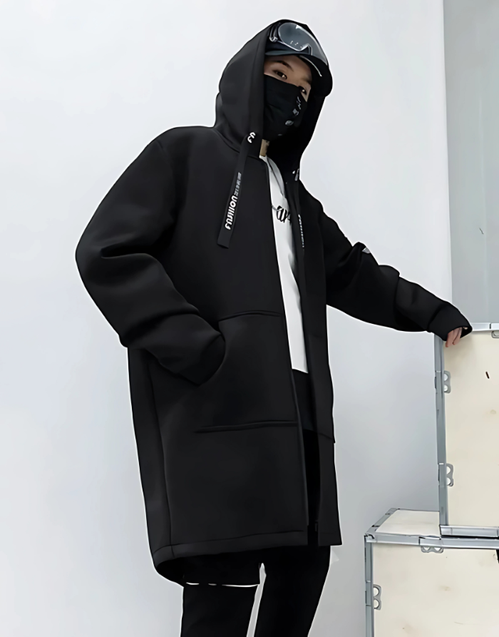 Person wearing a black hoodie with a brand logo, standing against a white wall.
