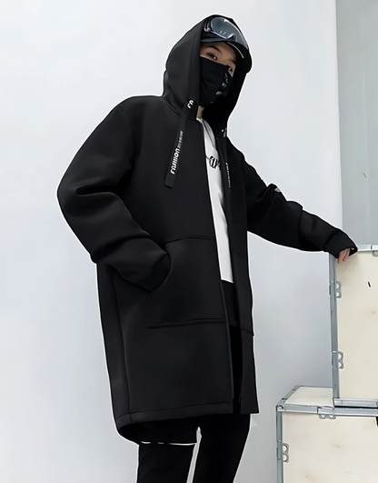 Person wearing a black hoodie with a brand logo, standing against a white wall.